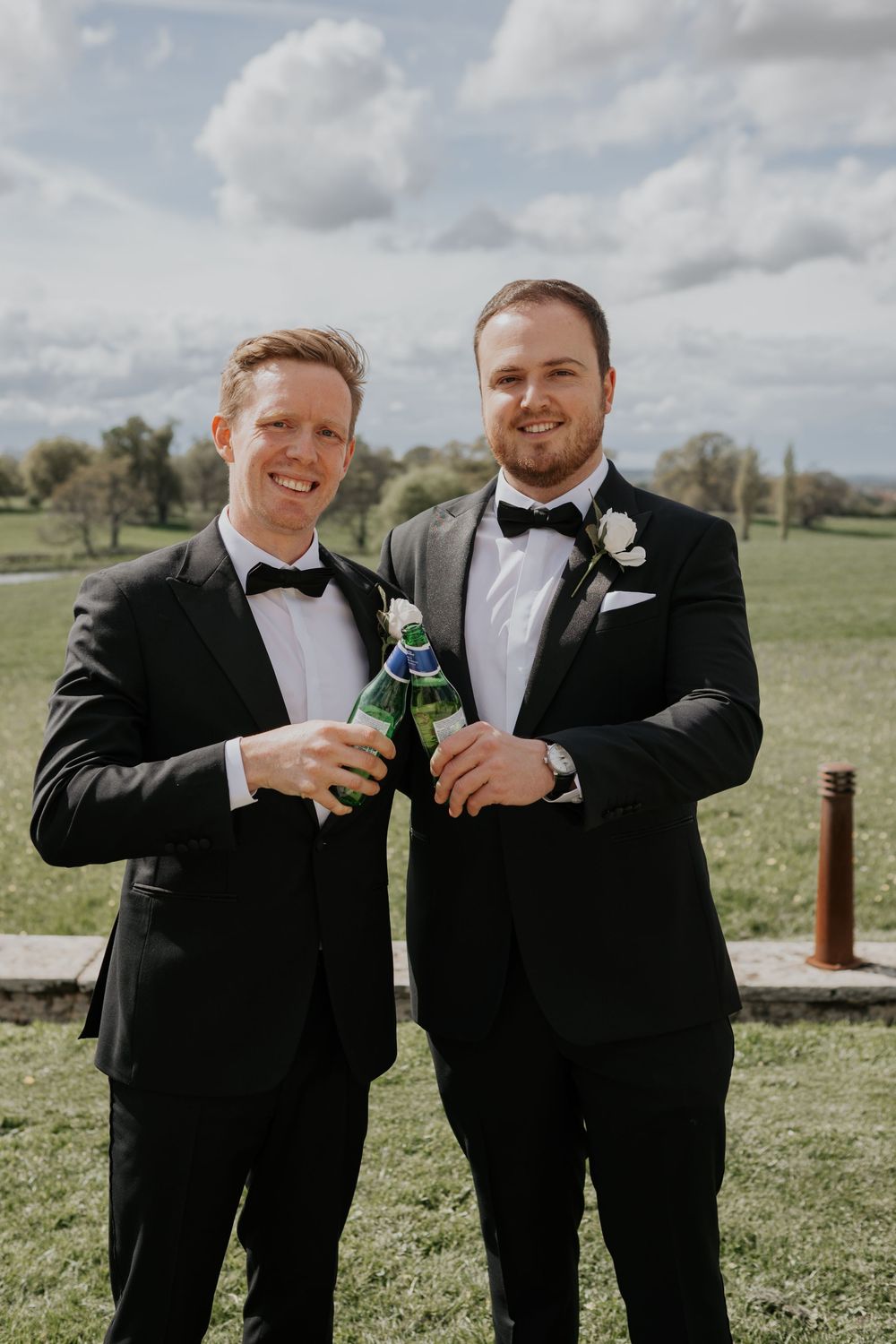 Two groomsmen in black tuxedos with bow ties toasting with beer bottles outdoors at a wedding.