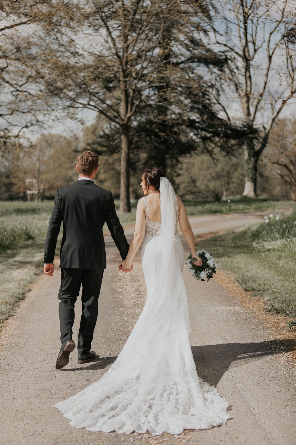 Bride in lace wedding gown with train and groom in dark suit walking hand in hand down a path.