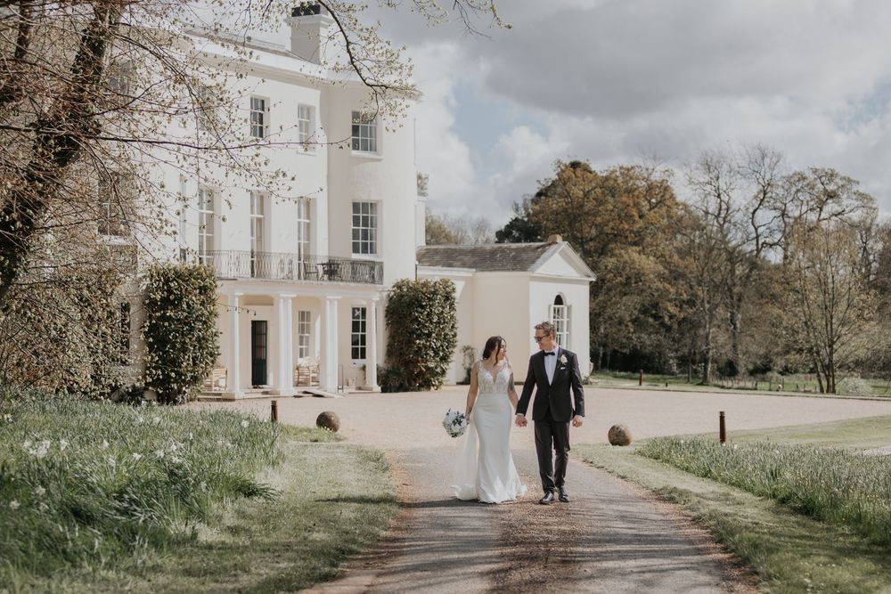 Bride and groom walking hand in hand along a path leading to a stunning white manor wedding venue.
