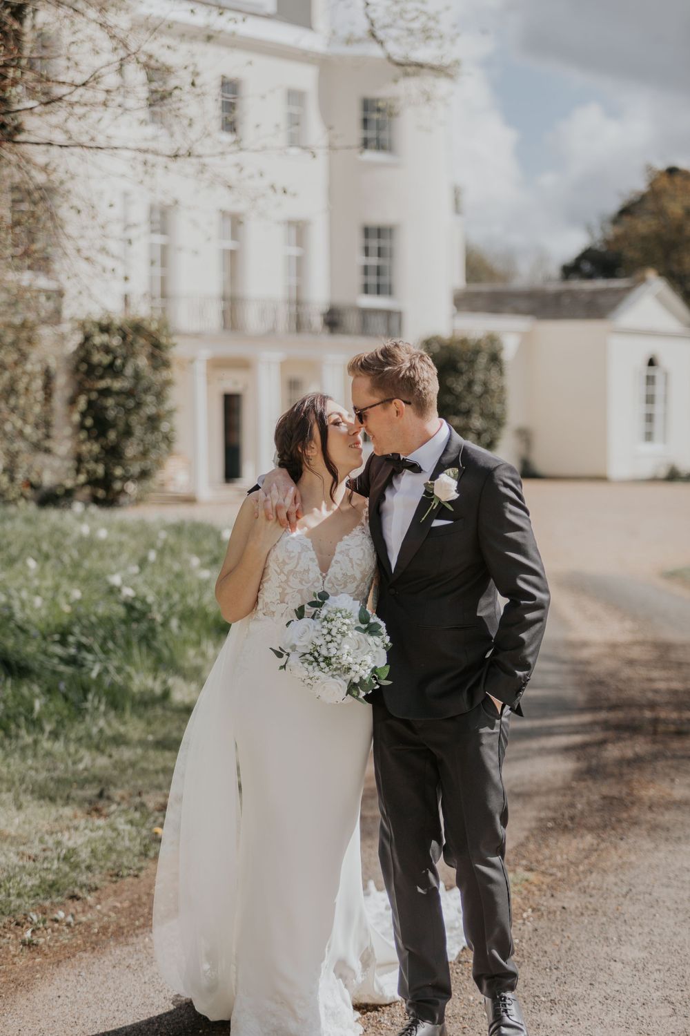 Bride holding white bouquet and groom in black tuxedo sharing a romantic moment outside a white manor.