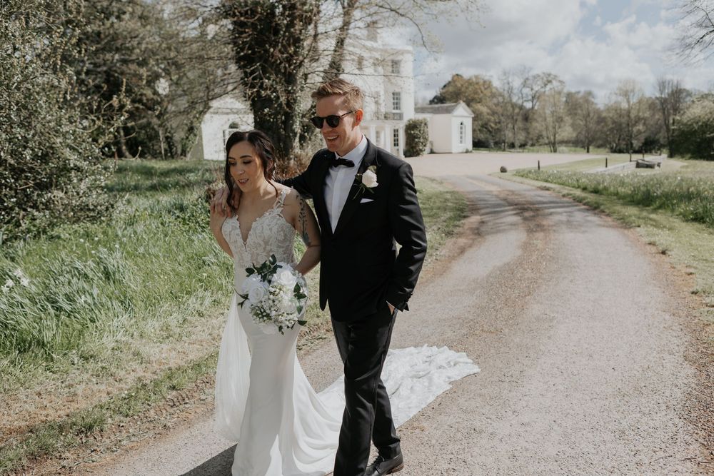 Bride in fitted lace gown and groom in black tuxedo walking together outside an elegant manor house.