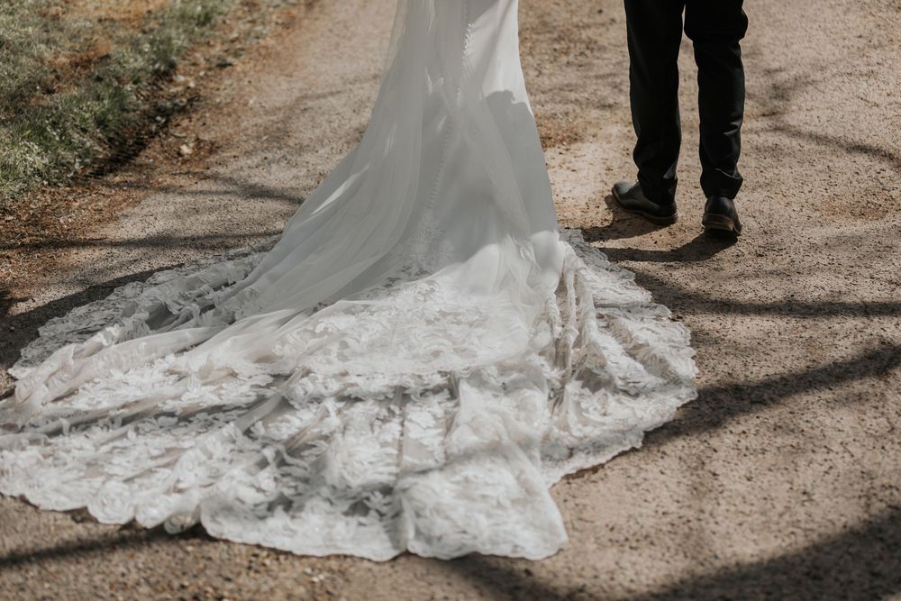 Close-up detail of a bride's intricate lace wedding gown train trailing on a gravel path.