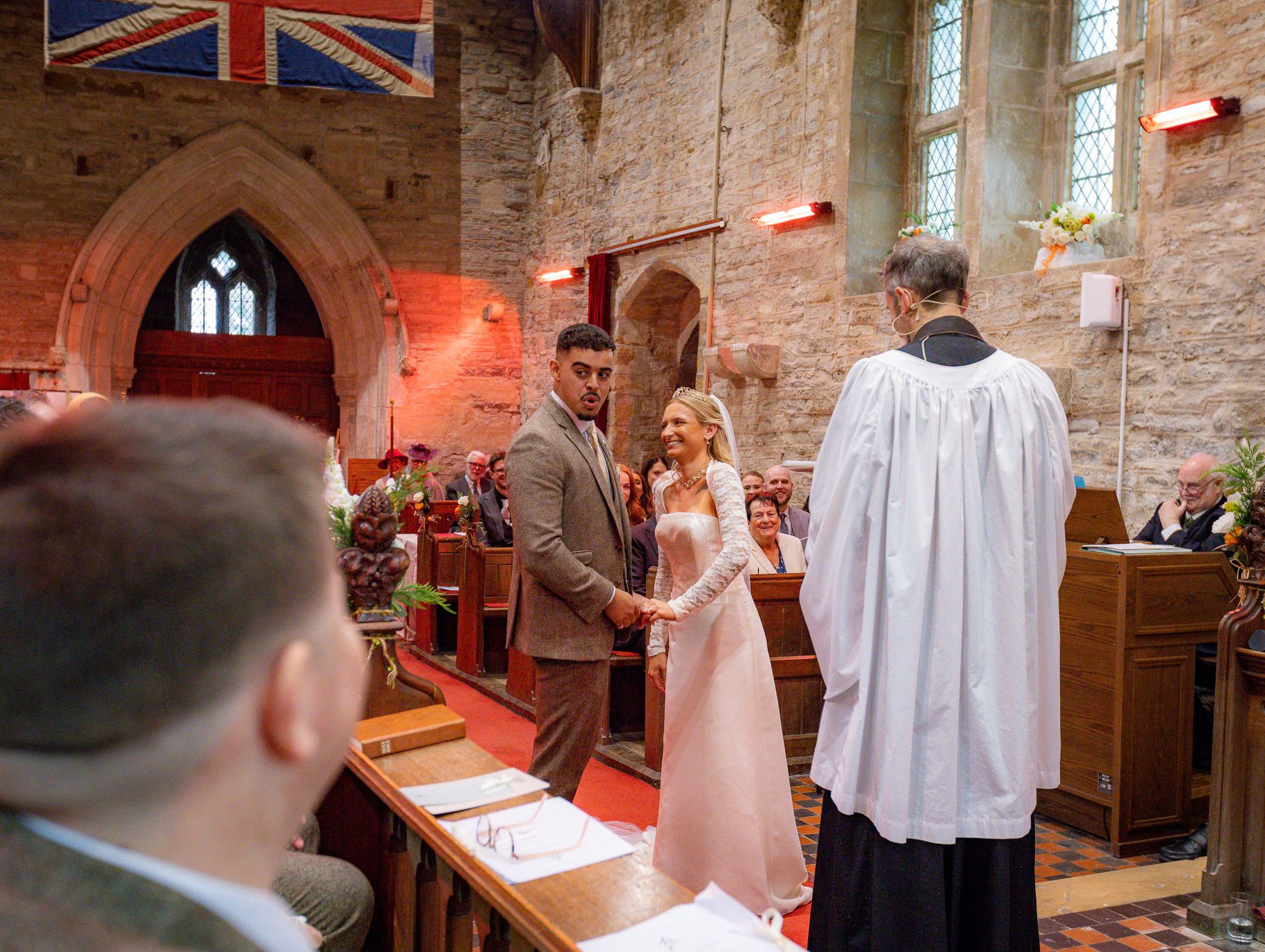 Wedding ceremony taking place inside a historic stone church with Union Jack flag visible in the background.