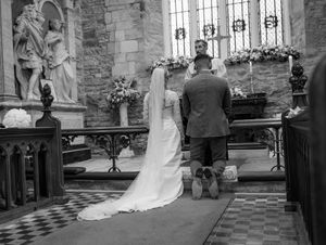 Black and white photograph of wedding ceremony in traditional church with checkered floor tiles.