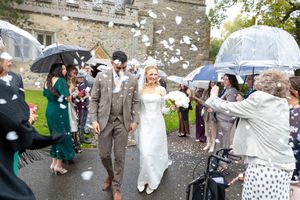 Wedding party celebrates outdoors with bubbles floating through the air as guests hold umbrellas on a cloudy day.