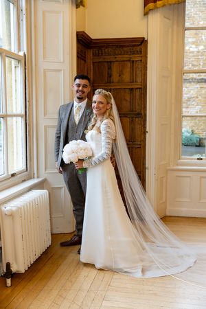 A newlywed couple poses in a sunlit room with wooden paneling and tall windows, the bride wearing a long-sleeved lace gown.