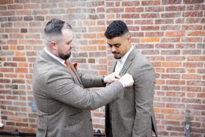Two men in grey suits help each other with boutonniere adjustments against a brick wall backdrop.