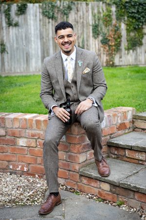 Person in a grey suit sitting on a brick wall outdoors with a garden backdrop.