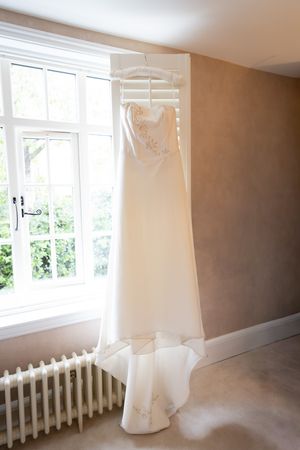 An elegant white wedding dress hangs on a decorative hanger near a bright window and radiator.