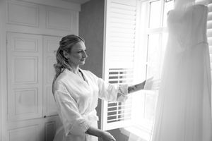 A person in a white robe examines a wedding dress hanging by a window in black and white.