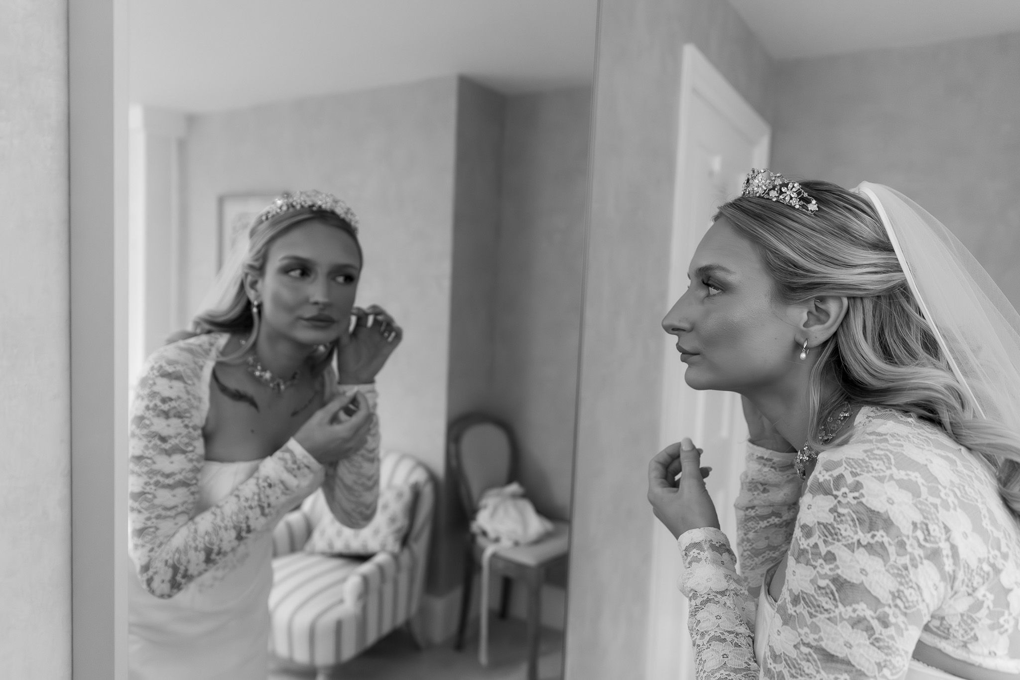 A bride in a lace dress applies lipstick while looking in a mirror during wedding preparations.