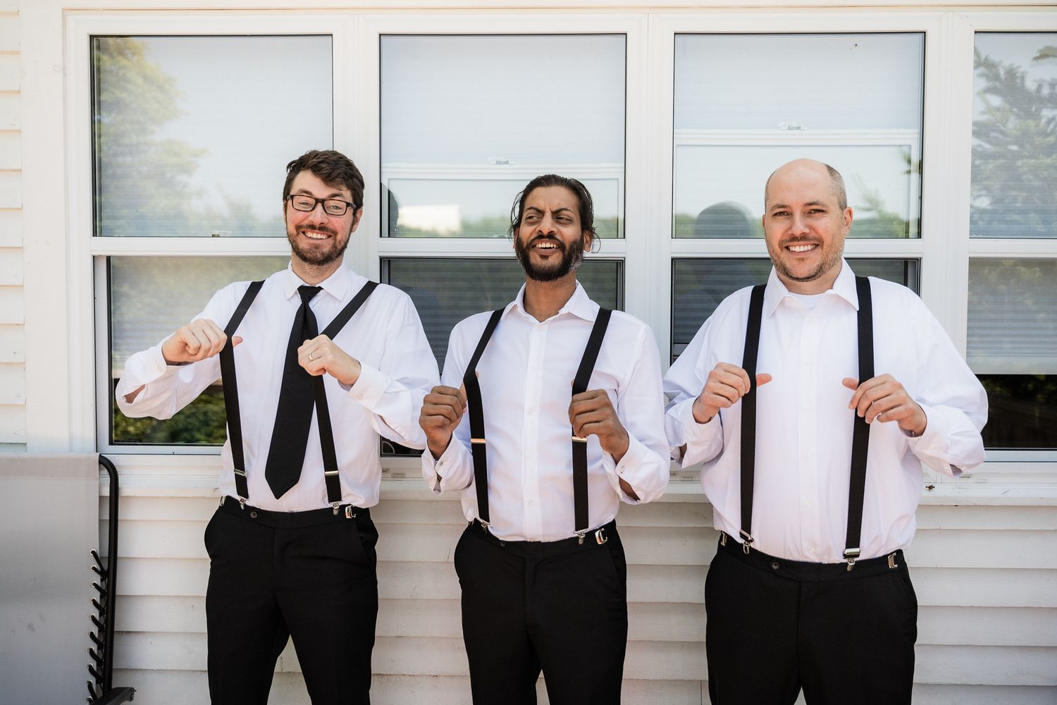 Groomsmen getting ready in front of a white house, one holding his suspenders and smiling, dressed in matching black-and-white outfits.