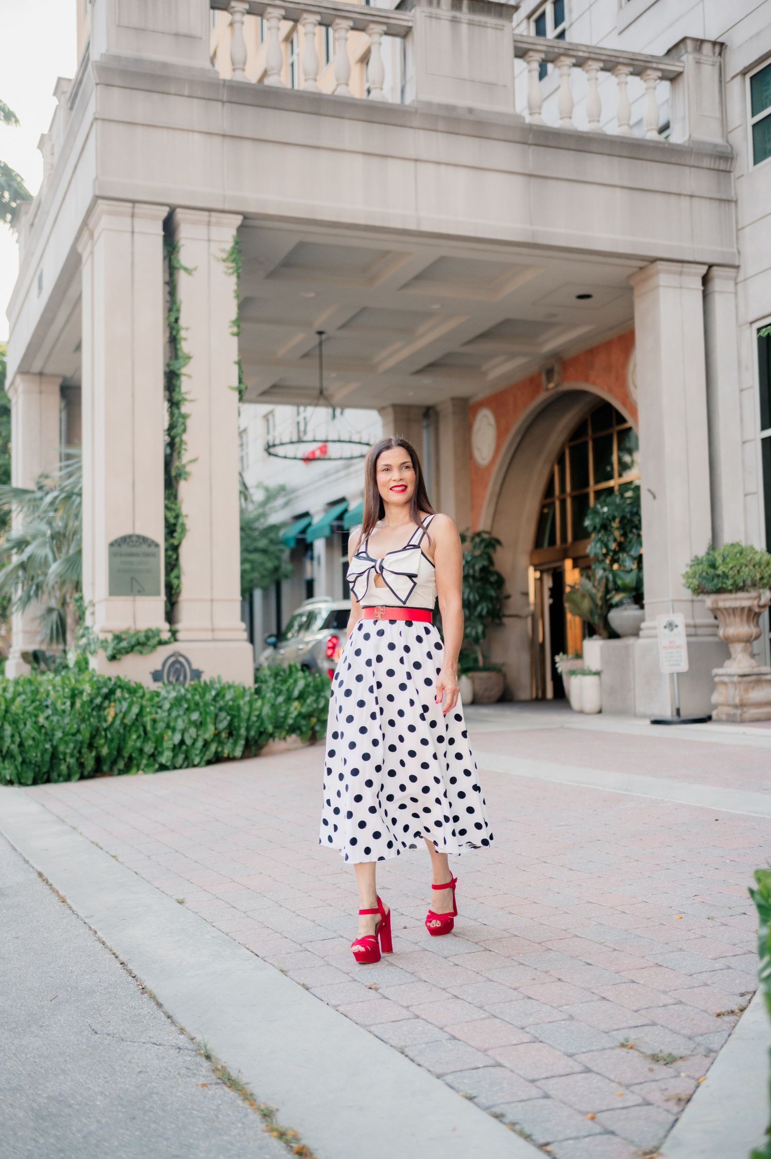 A polka dot dress and red shoes create a stylish look against a stone architectural backdrop.
