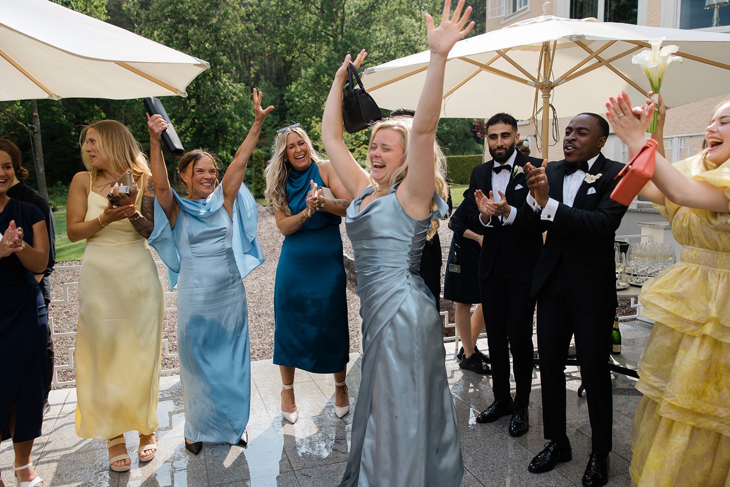 Wedding party celebrates joyfully under white umbrellas with groomsmen in black tuxedos and bridesmaids in colorful dresses.