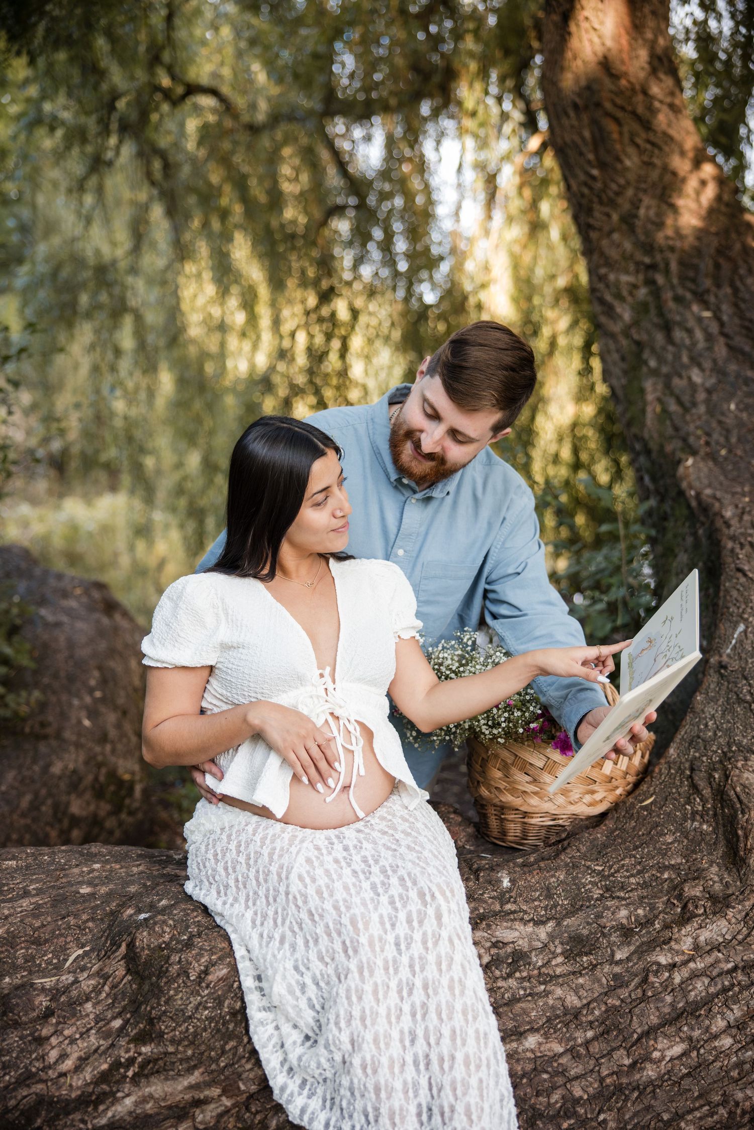 A couple in a white dress and blue shirt share an intimate moment reading together under a tree in nature.