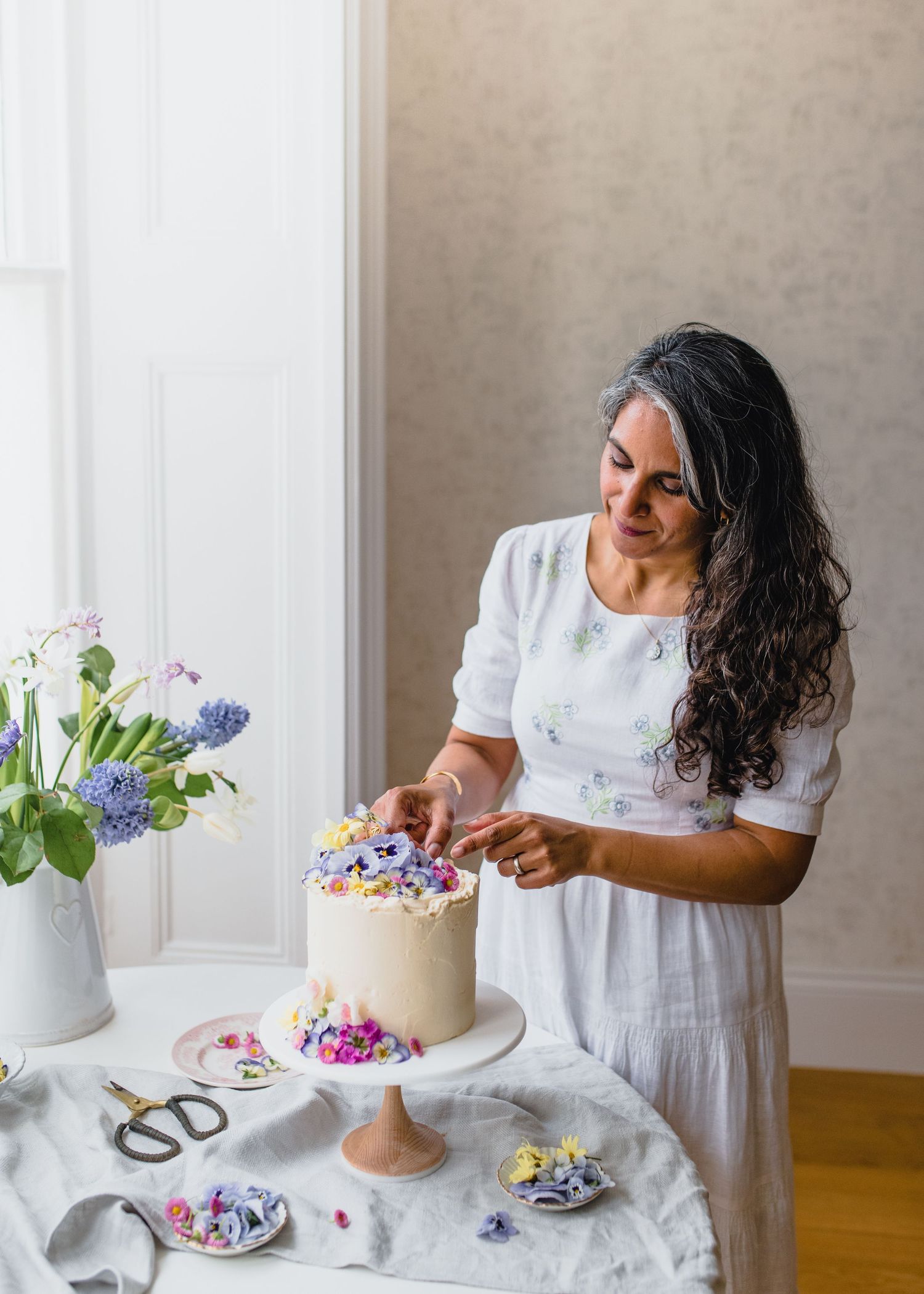 Baker in white dress decorates tiered cake with fresh flowers and delicate piping details on wooden table.