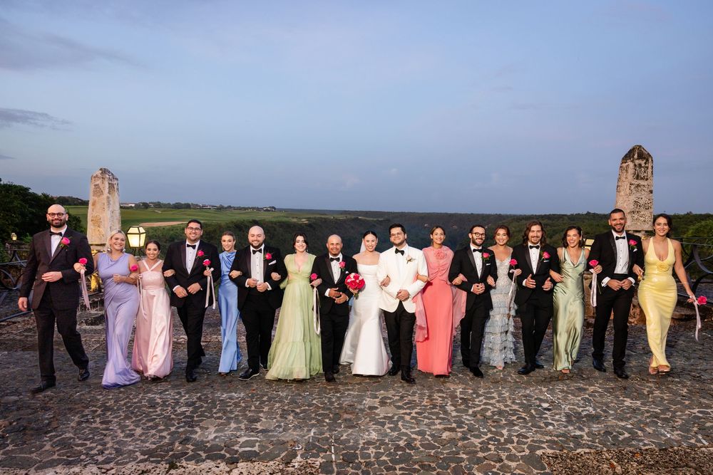 A wedding party stands in a line at sunset on a stone patio with ornate towers in the background.