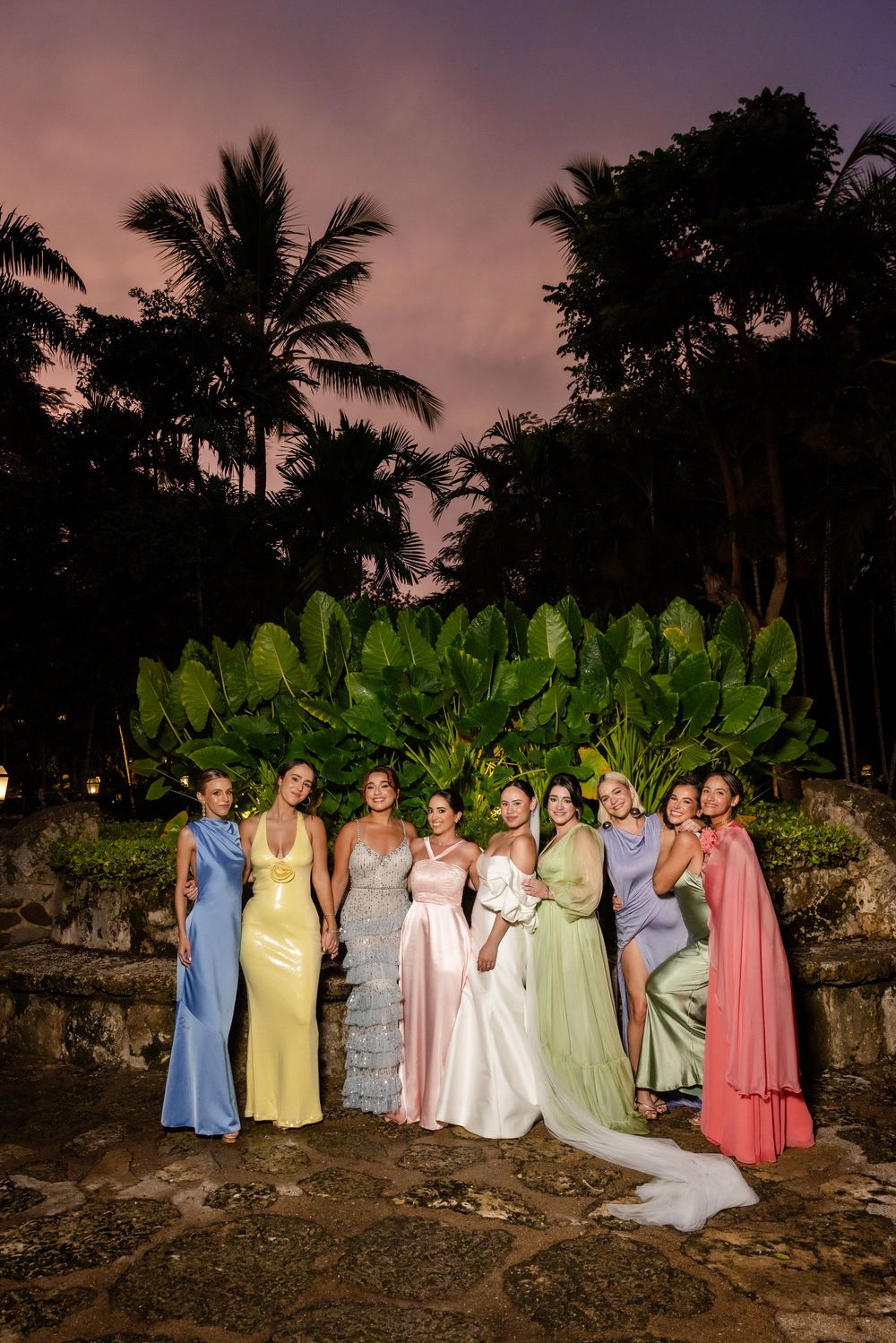 A wedding party in pastel colored dresses poses at sunset with palm trees and tropical foliage in the background.