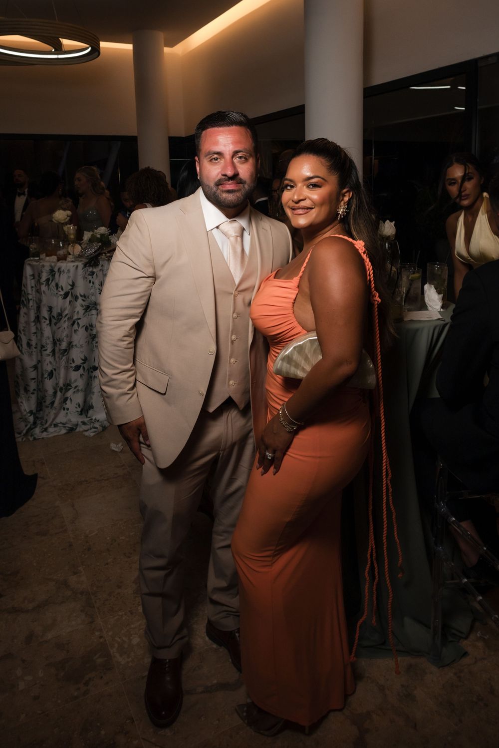 A couple in formal attire, with a person wearing a beige suit and another in a coral-colored dress, posing at an event.