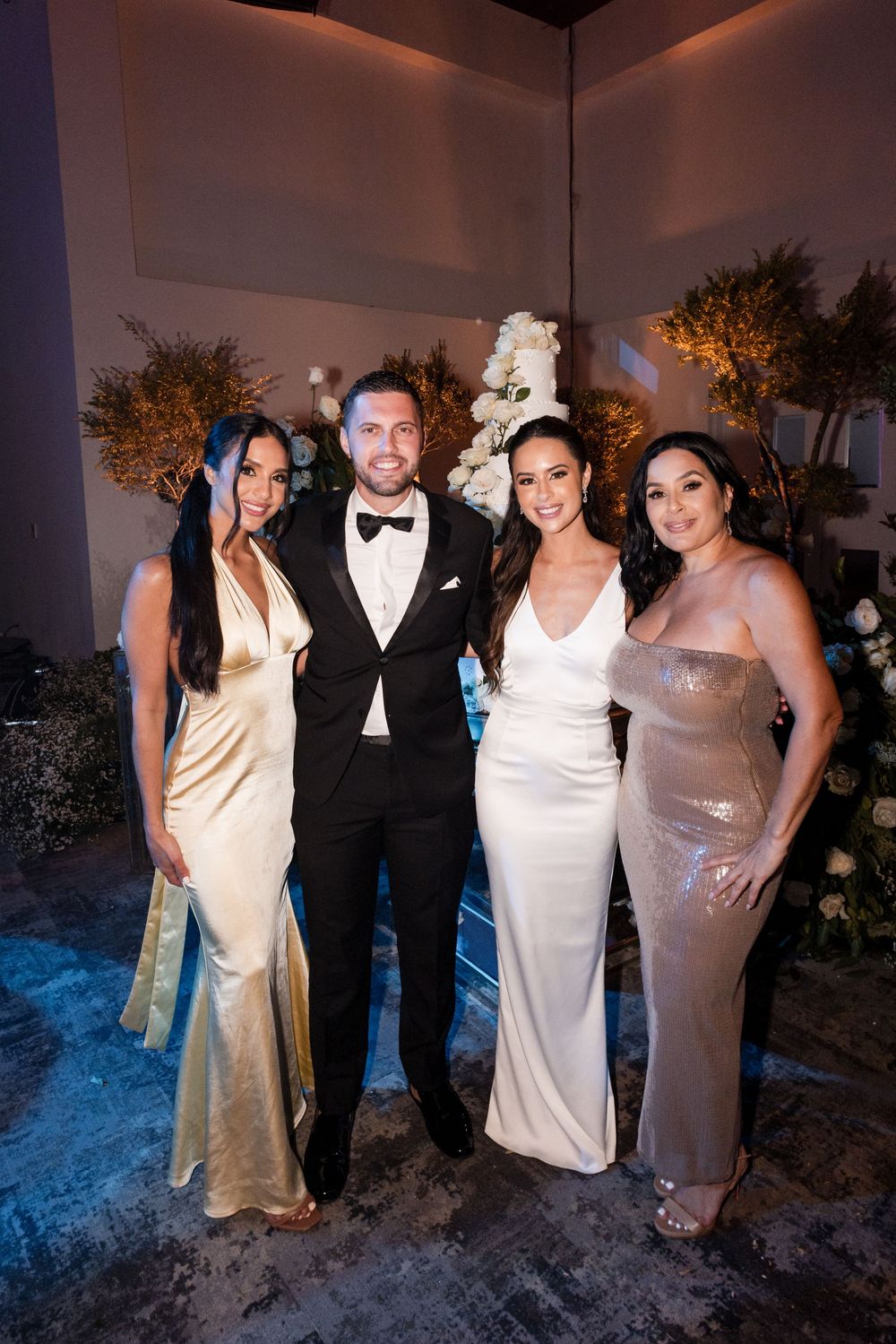 Wedding party members gather for photos wearing formal attire in neutral tones at an evening reception.