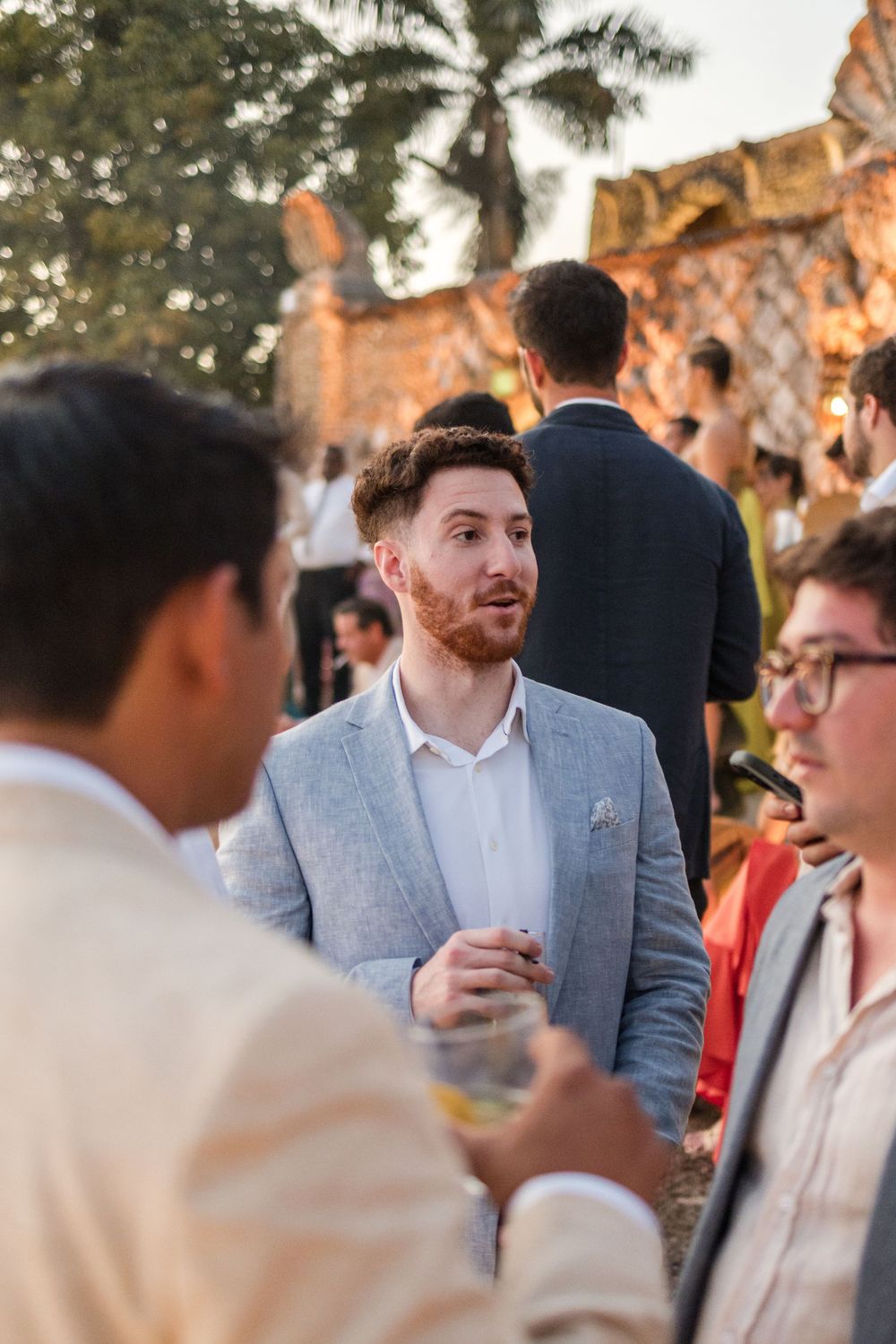 A group of guests having a conversation at an outdoor event with palm trees and stone architecture in the background.
