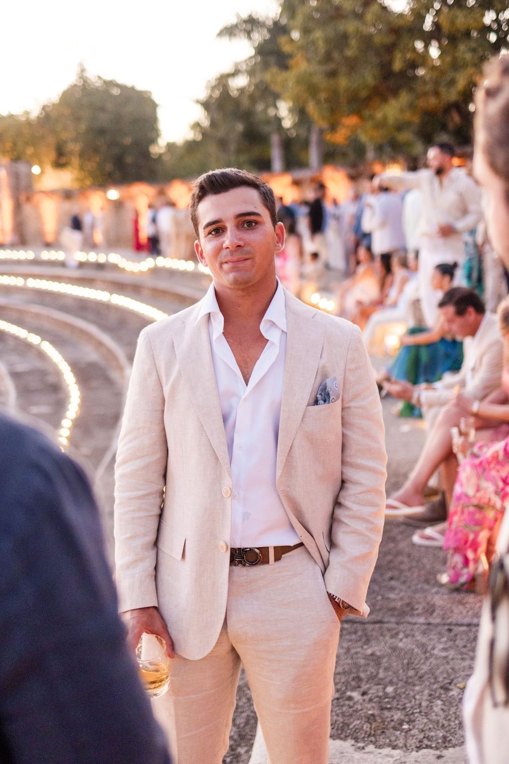 A person in a beige linen suit and white shirt stands at an outdoor event during golden hour.