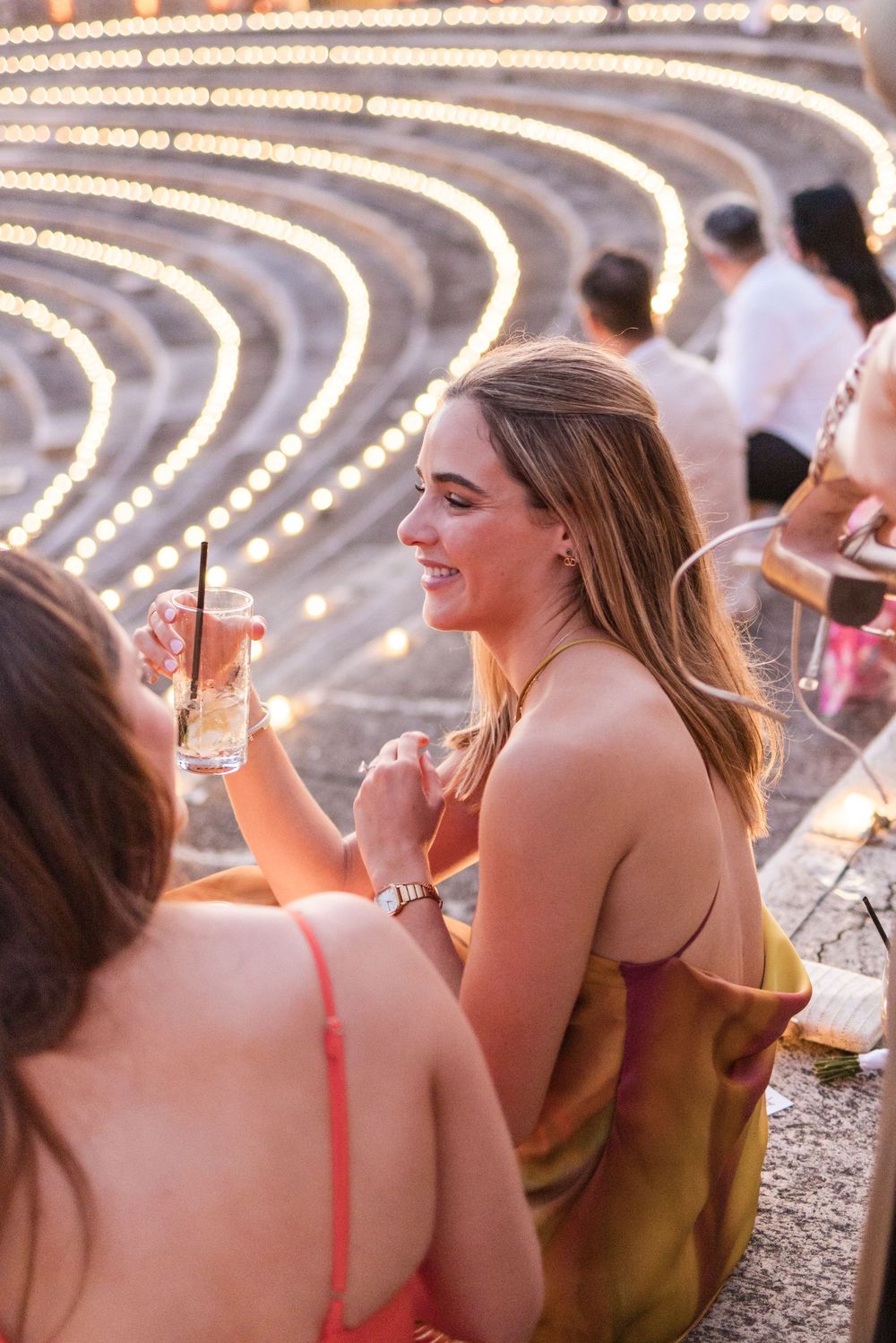 People enjoying drinks and conversation at an outdoor event with curved string lights overhead.