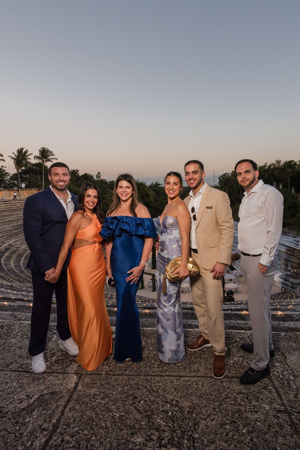 Group photo of wedding attendees posing together on a beach at dusk wearing formal attire.