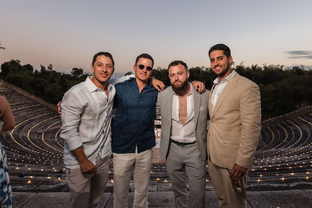 Four friends pose together at an outdoor amphitheater during sunset wearing casual formal attire.