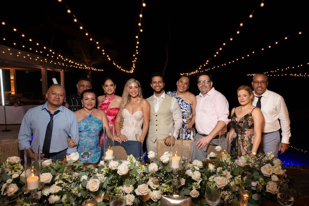 Wedding party poses behind elegant floral table arrangement under romantic string lights at evening reception.