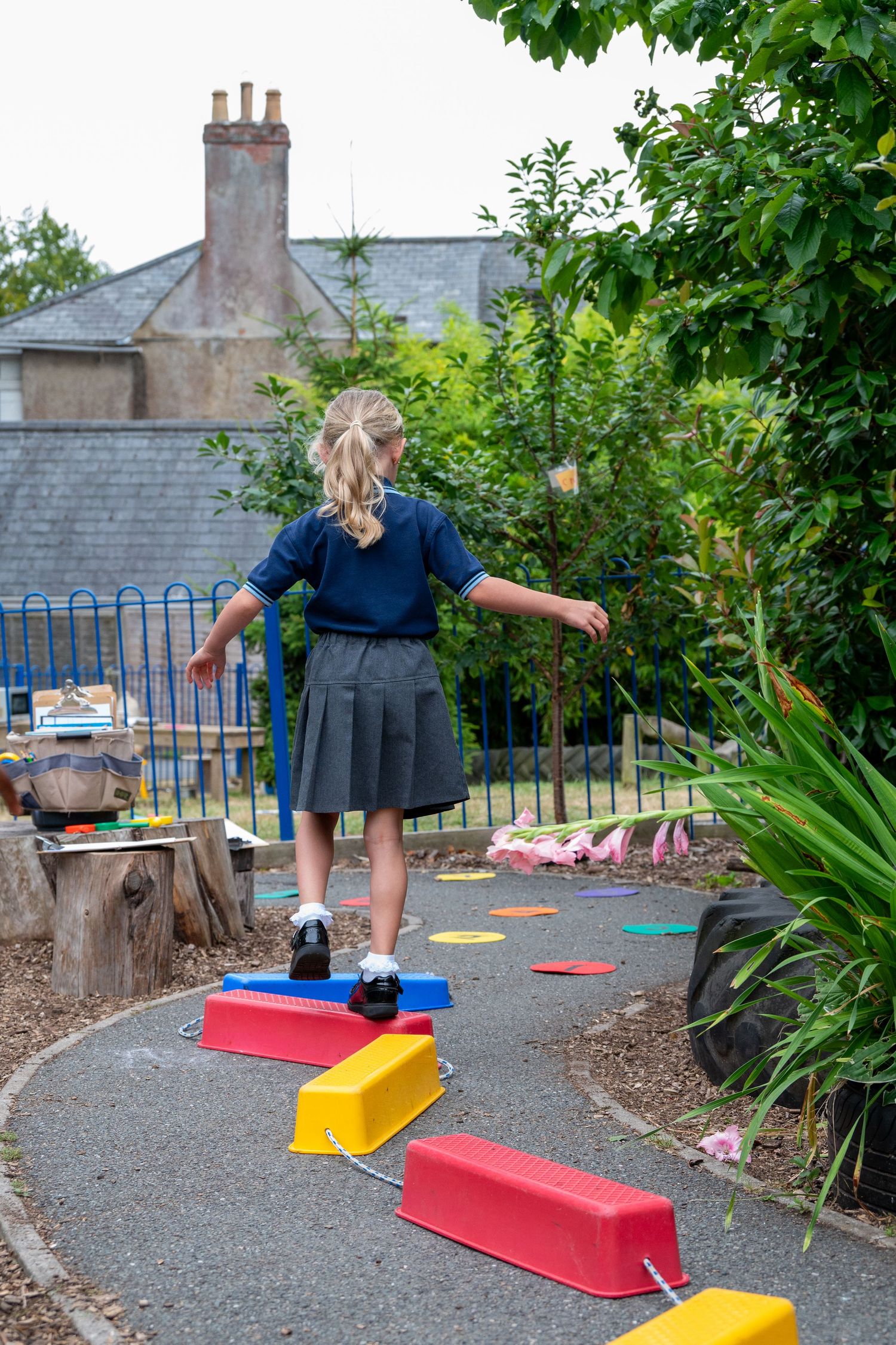 A child in school uniform balances on colorful outdoor play equipment in a gravel playground area.