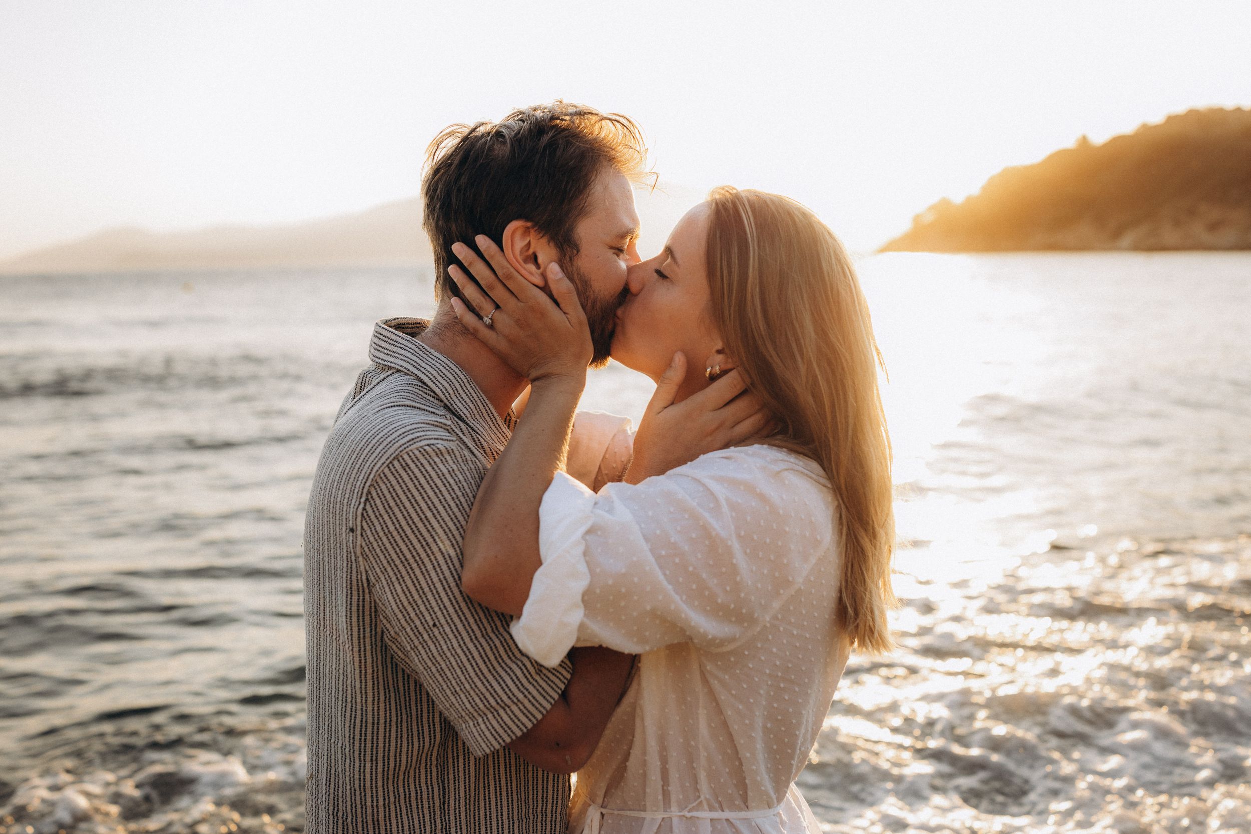 A couple shares romantic kisses by the ocean during golden hour at sunset.