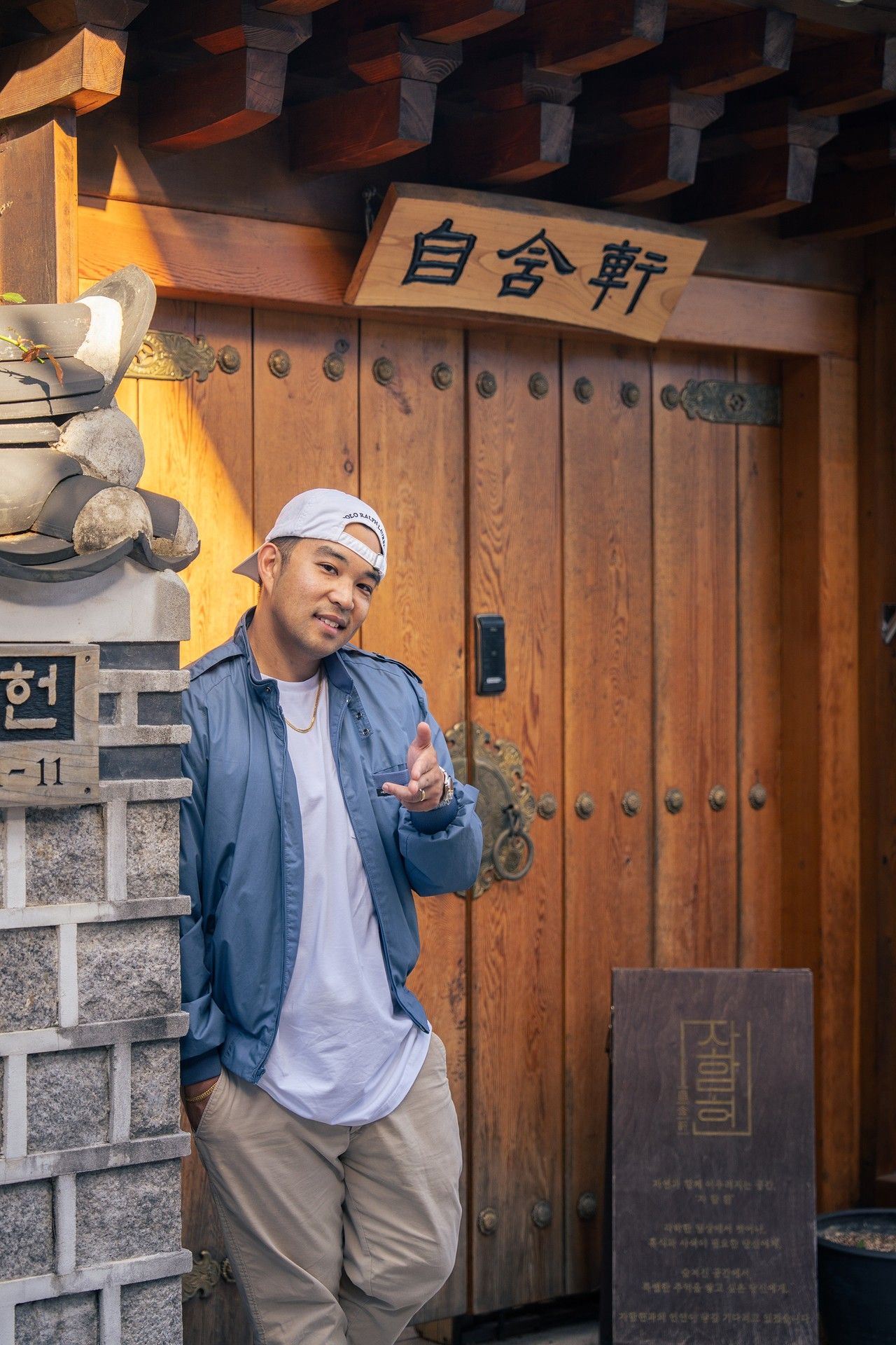 A person in casual attire poses in front of a traditional wooden door with Korean text and stone walls.