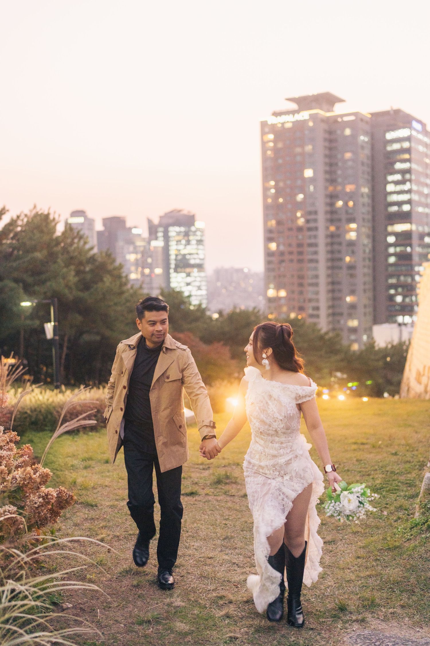 A couple walks through a scenic outdoor setting at sunset with city buildings in the background.