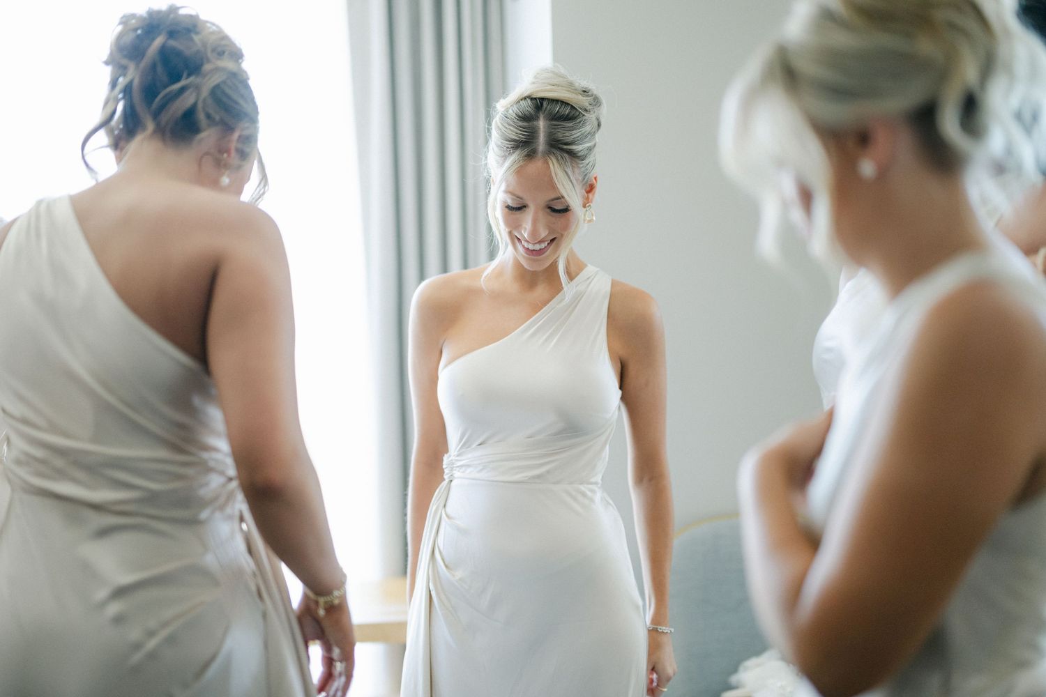 Elegant one-shoulder white wedding dress being fitted in a bright room with natural lighting.