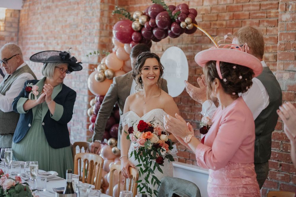 Smiling bride walks through reception room as guests in hats applaud near burgundy balloon arch.