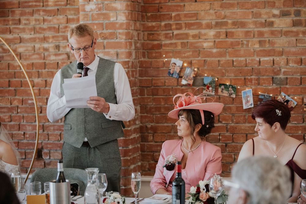 Man in grey waistcoat delivers wedding speech at microphone while guests in hats listen attentively.