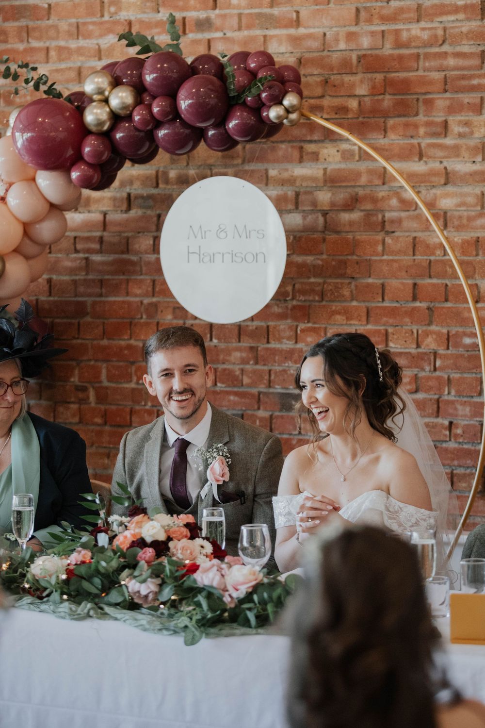 Mr and Mrs Harrison smile at head table beneath burgundy and gold balloon arch at wedding reception.