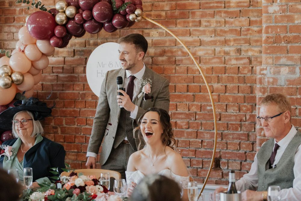 Groom speaks into microphone at wedding reception as bride laughs beside balloon arch backdrop.