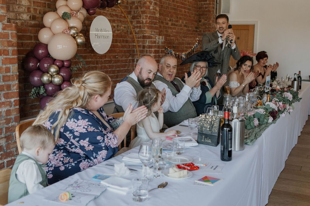 Wedding party claps and celebrates together at decorated head table during lively reception dinner.