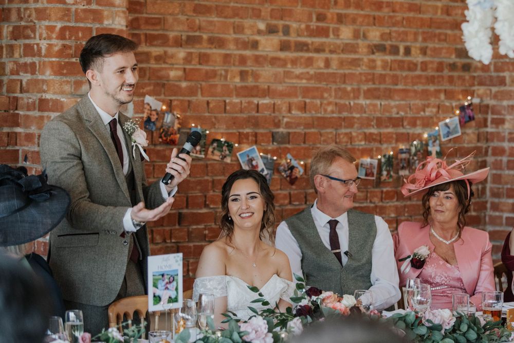 Young groomsman delivers wedding speech as bride and guests smile at floral decorated reception table.