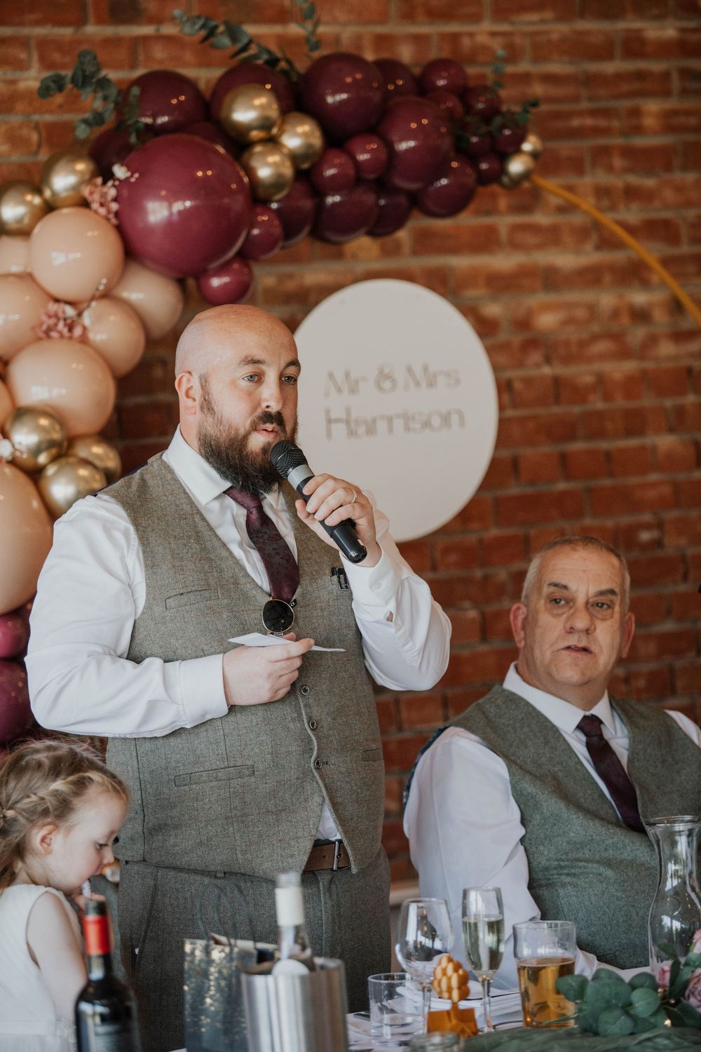 Bald bearded man gives wedding speech at microphone beside Mr and Mrs Harrison balloon backdrop.