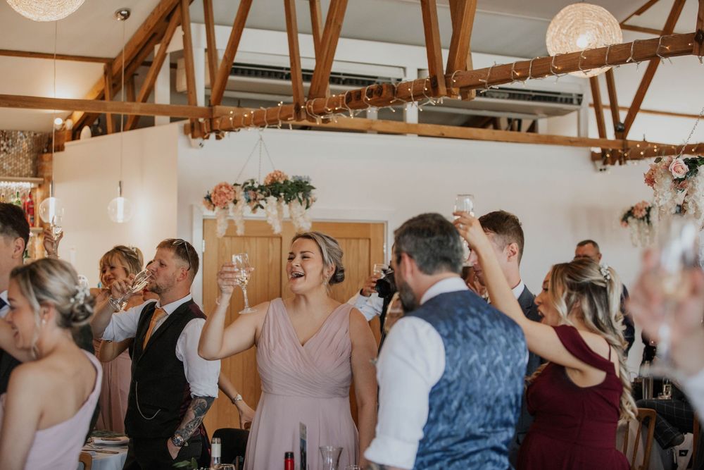 Wedding guests celebrate and raise glasses in a rustic barn reception venue decorated with fairy lights and floral arrangements.