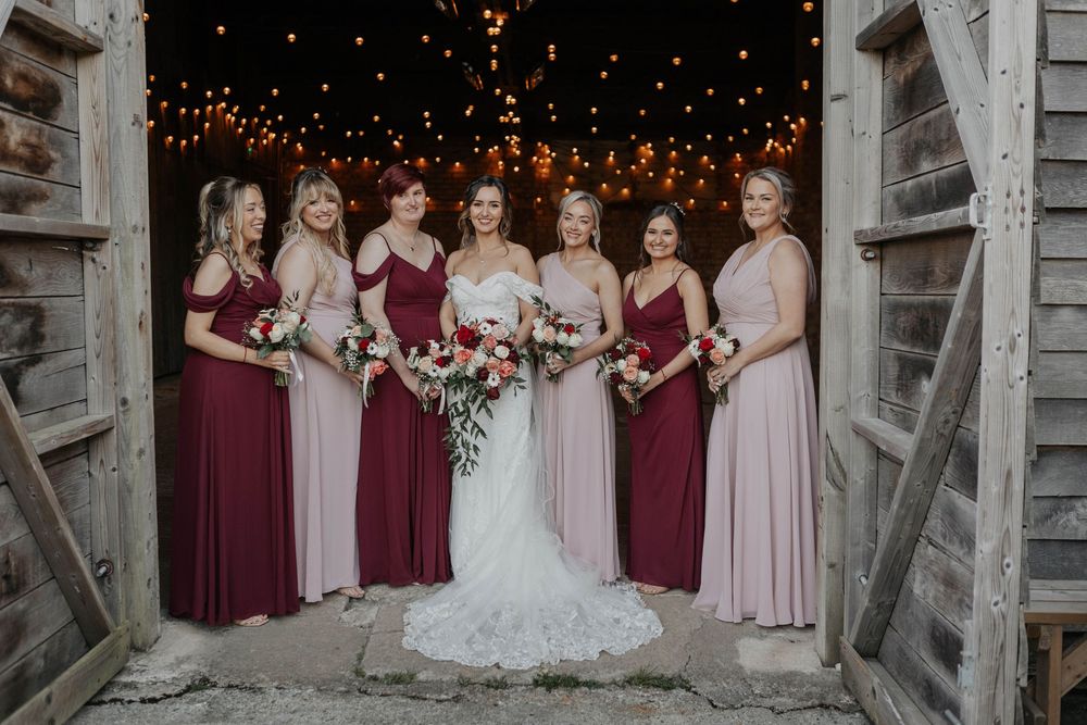 Bride in lace gown poses with six bridesmaids in burgundy and blush dresses inside a rustic barn with fairy lights.