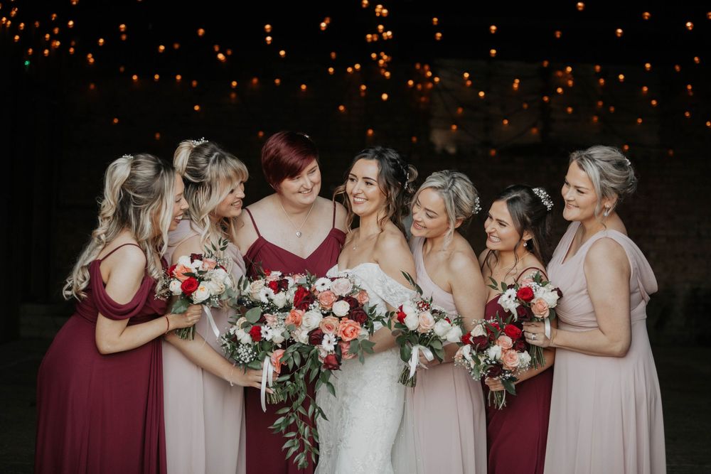 Bride and bridesmaids in burgundy and blush gowns laugh together while holding bouquets of red and pink flowers outdoors.