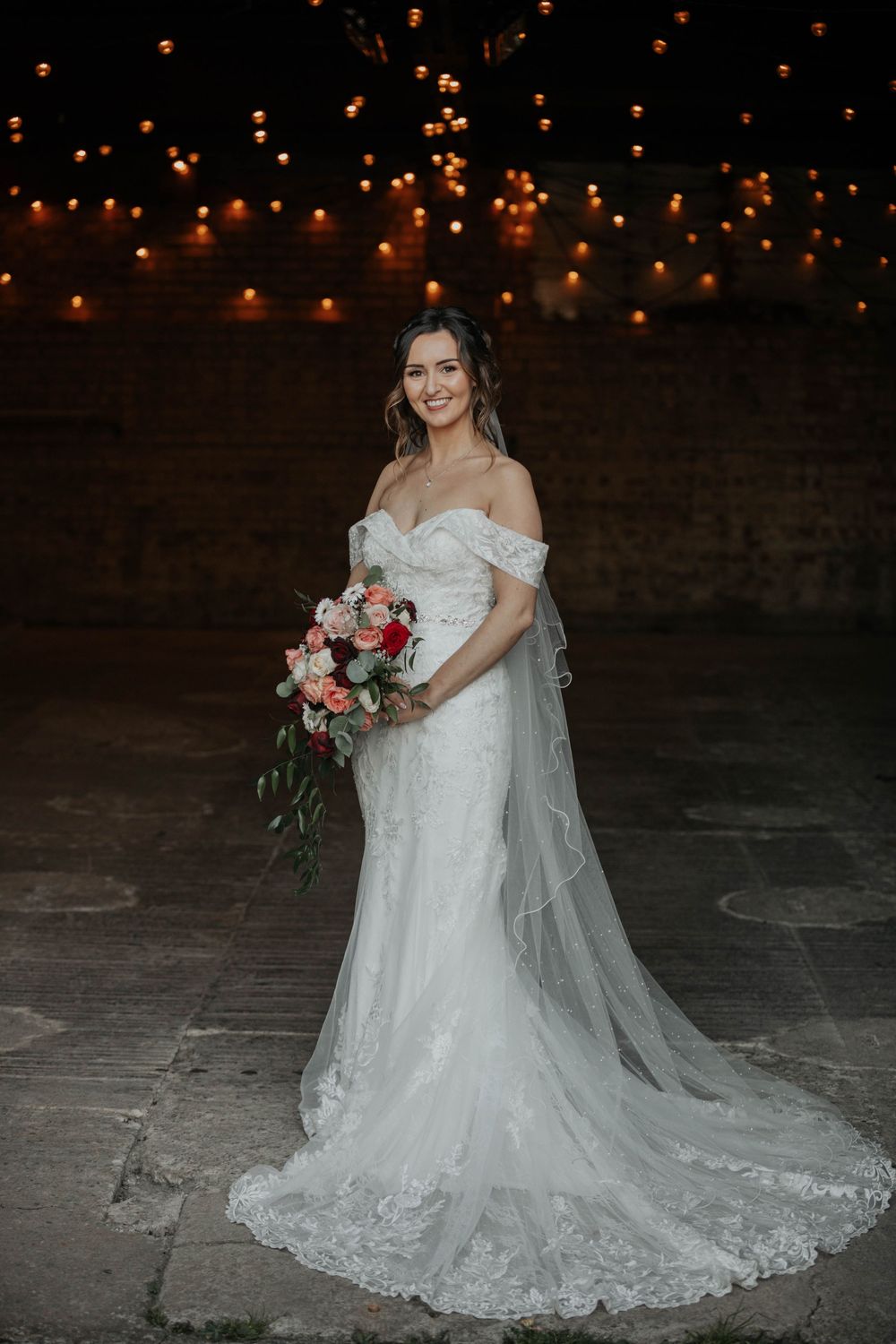Elegant bride in off-shoulder lace wedding gown holds a cascading bouquet of red and pink roses under warm fairy lights.