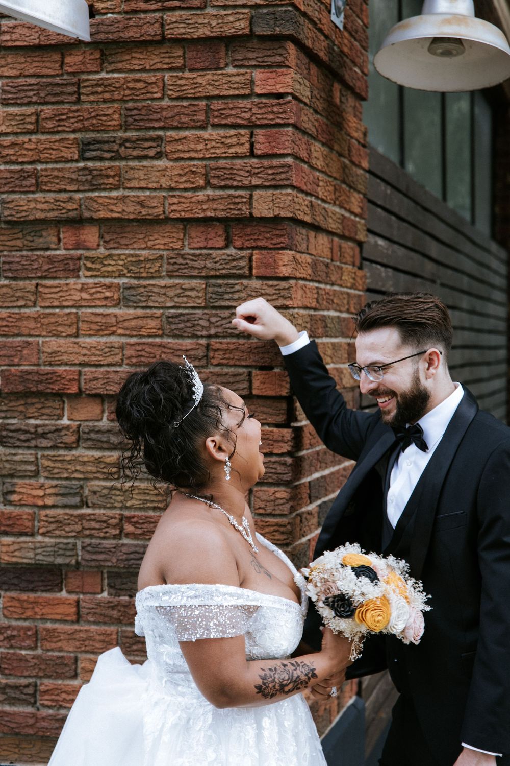 A joyful wedding moment shared between a couple against a brick wall background with a white bouquet of flowers.