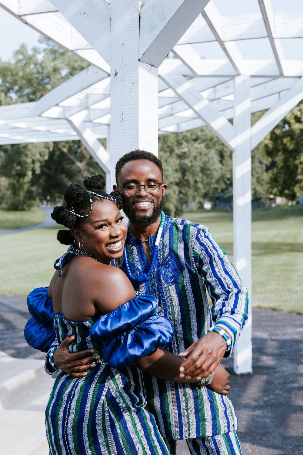 A couple in matching blue and white striped outfits embrace and laugh together under a white pergola structure.