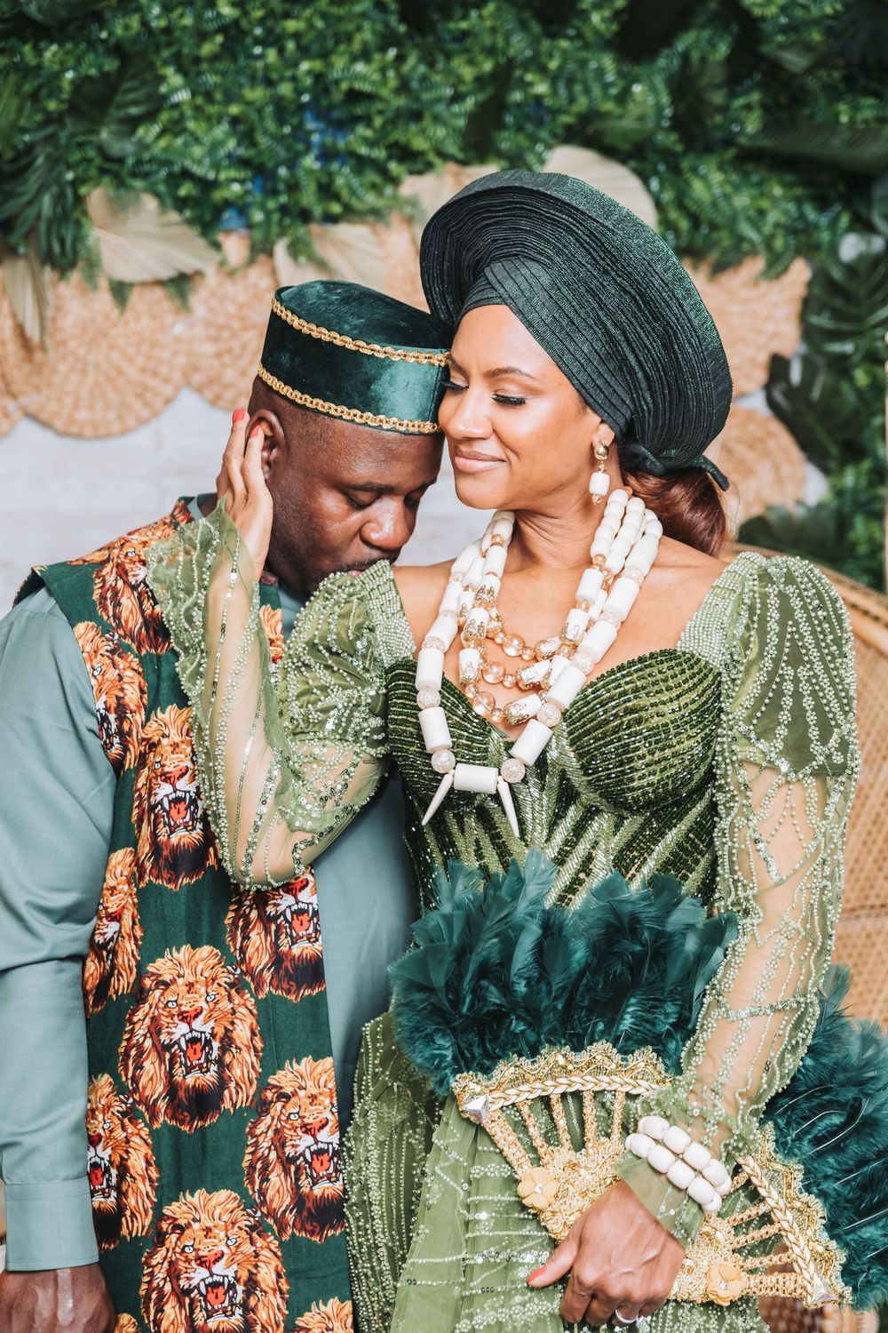 A couple in traditional African attire sharing romantic moments outdoors against a lush green background.