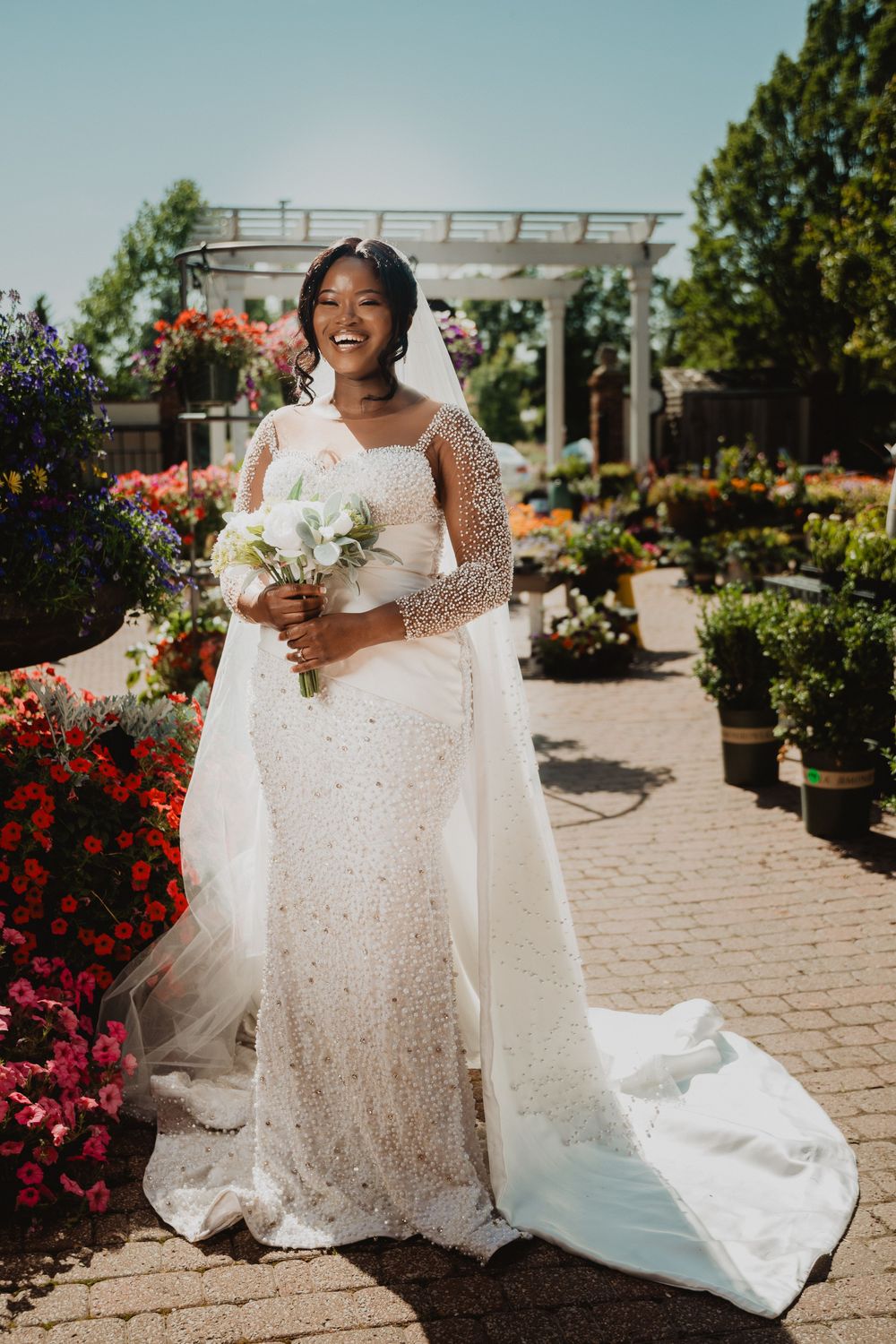 A beautiful bride in a sparkling white lace gown poses in a garden setting with colorful flowers and stone pathways.